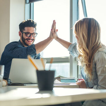 Zwei Personen an einem Tisch klatschen sich mit den Händen ab. High Five.