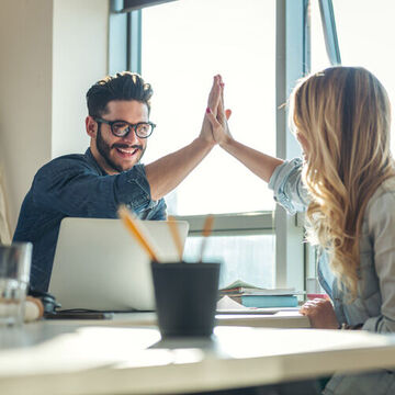 Zwei Personen an einem Tisch klatschen sich mit den Händen ab. High Five.