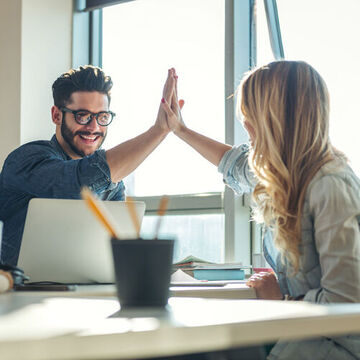 Zwei Personen an einem Tisch klatschen sich mit den Händen ab. High Five.
