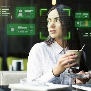 Eine Frau sitzt mit einem Getränk vor einem Laptop, während digitale Overlays Gesichtserkennung und Datenanalyse symbolisieren. Das Bild veranschaulicht den Einsatz von Künstlicher Intelligenz (KI) und biometrischen Technologien zur Identifizierung und Verarbeitung personenbezogener Informationen im digitalen Umfeld.