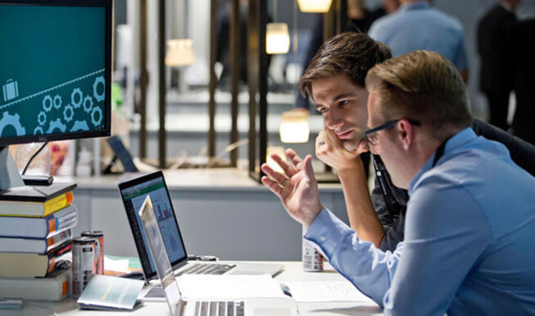 Bild von zwei Personen, die am Schreibtisch sitzen, auf einen Computer schauen und diskutieren.