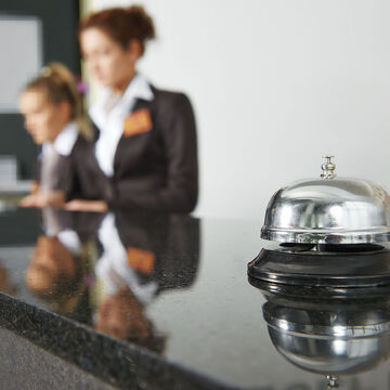 Bild von einer Hotelrezeption. Im Vordergrund steht eine Klingel auf dem Tresen und im Hintergrund befinden sich zwei Personen.