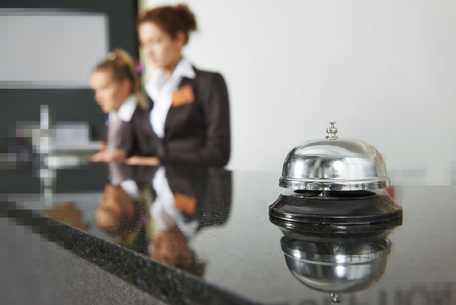 Bild von einer Hotelrezeption. Im Vordergrund steht eine Klingel auf dem Tresen und im Hintergrund befinden sich zwei Personen.
