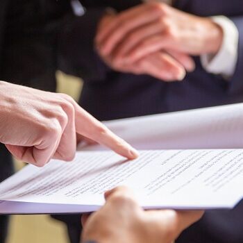 Mehrere Personen in Anzügen stehen zusammen und betrachten ein Dokument. Eine Person zeigt mit dem Finger auf eine Textstelle, während eine andere das Papier hält