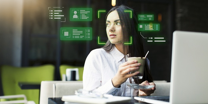 Eine Frau sitzt mit einem Getränk vor einem Laptop, während digitale Overlays Gesichtserkennung und Datenanalyse symbolisieren. Das Bild veranschaulicht den Einsatz von Künstlicher Intelligenz (KI) und biometrischen Technologien zur Identifizierung und Verarbeitung personenbezogener Informationen im digitalen Umfeld.